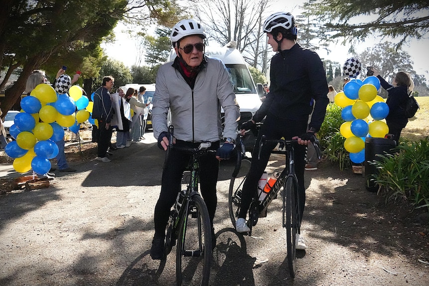 Two men on bikes with van behind them; blue and yellow balloons on either side of road.