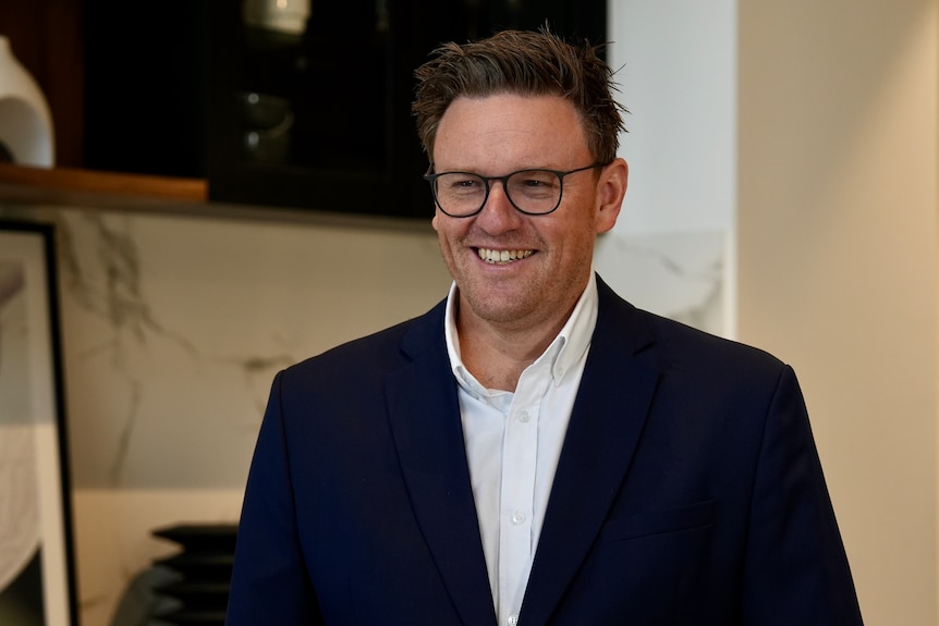 A man wearing a navy suit and white business shirt and glasses smiles while standing in the kitchen of a display home.