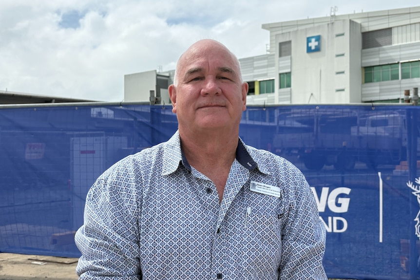 A middle-aged man in a collared shirt stands in front of a fenced-off construction site in front of a multi-storey hospital.