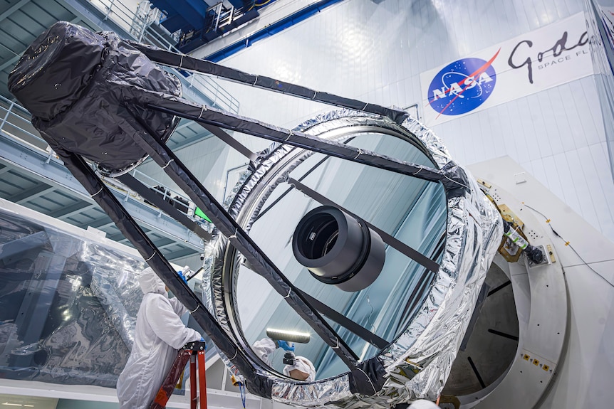 A person in white coveralls and a face mask stands on a ladder and shines a light at a massive round mirror in a NASA warehouse.
