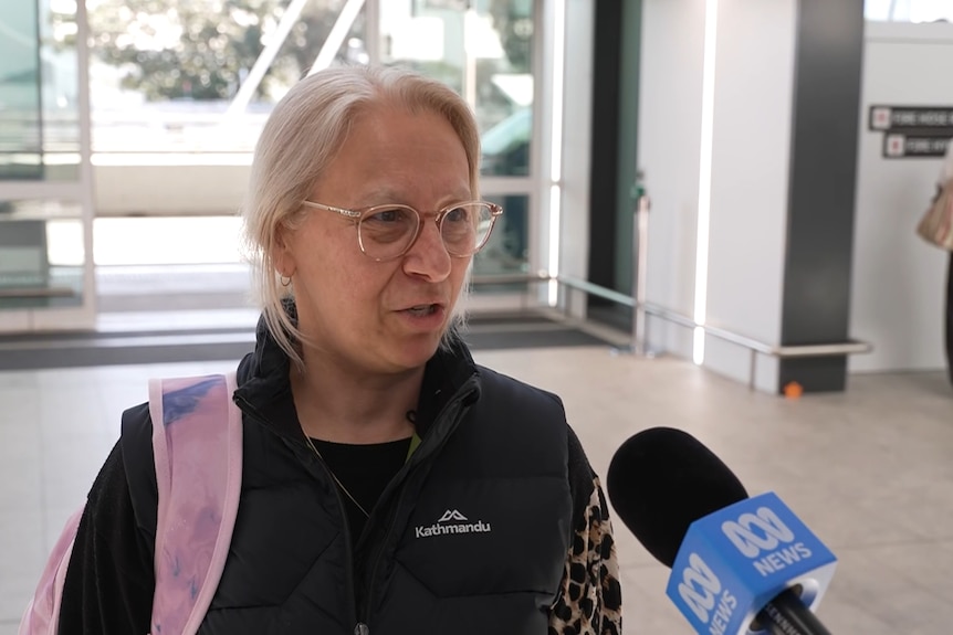 Louise with grey hair, glasses, in a Kathmandu black puffer jumper, speaking into an ABC microphone at the airport.