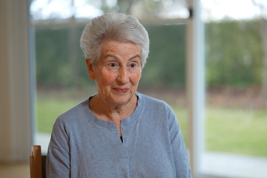 Nina is an elderly woman with short light-coloured hair. She has a neutral expression and dark clothes.