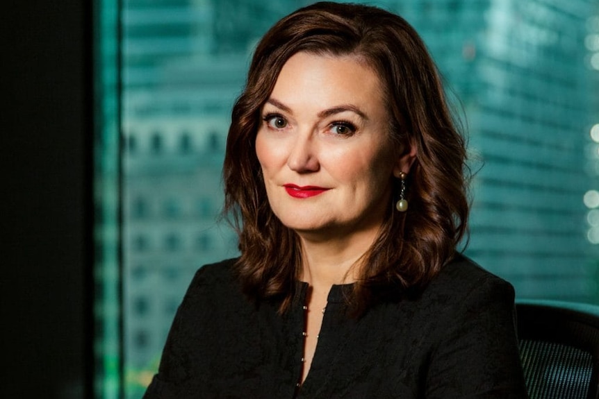 A woman in a black business shirt and red lipstick sits in an office in front of highrise buildings.