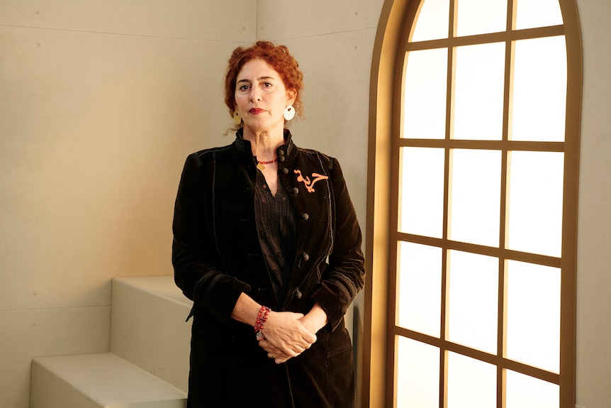 A woman with red hair in a black dress stands in front of a window