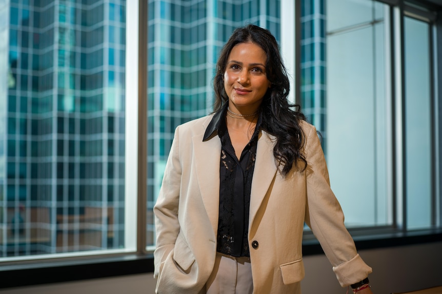 Esha Oberoi stands at a window, with tall building behind her
