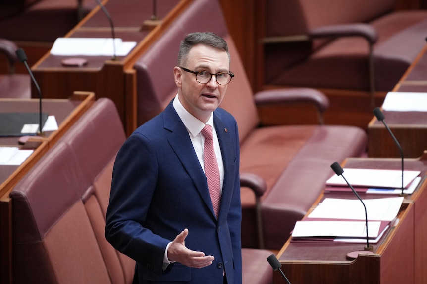 A white man with short brown hair and glasses wearing a dark blue suit stands in the Senate Chamber