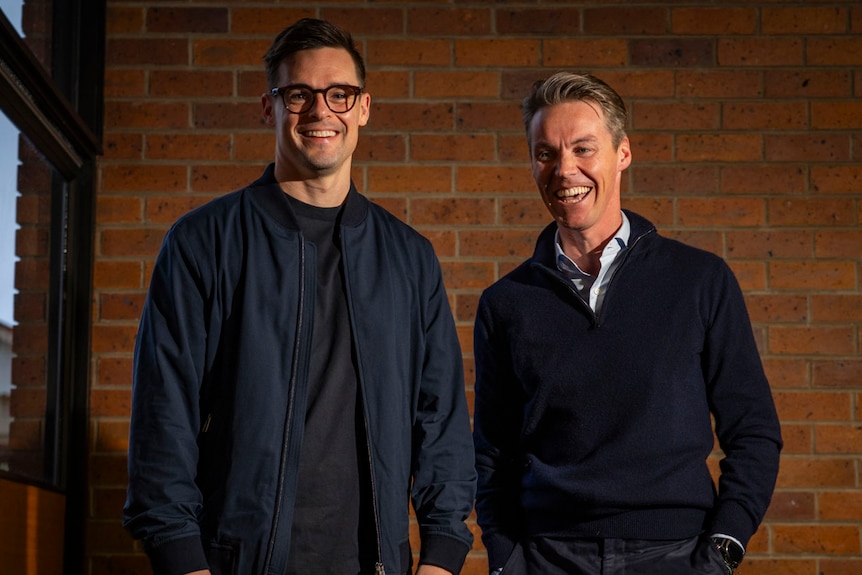 two men in dark tops stand against a red brick wall, one brunette and taller, the other with grey hair