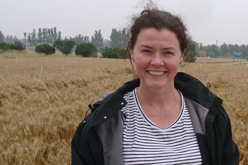A woman, wearing a jacket, standing in a field while smiling at the camera.