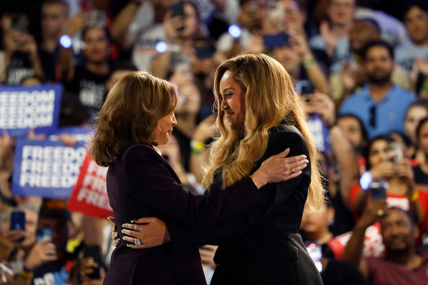 Two women with brown hair hug wearing black blazers.