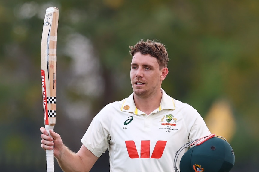 A cricketer in white celebrates a century, holding his bat and helmet