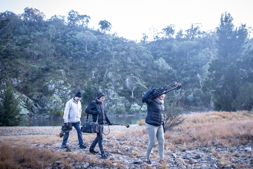 A woman and two men wear long pants and puffer jackets while carrying camera gear in the bush.