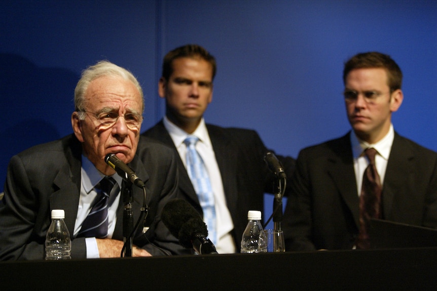 Rupert, Lachlan, James Murdoch sit at a press conference table wearing suit and ties. Rupert at centre in front of microphone