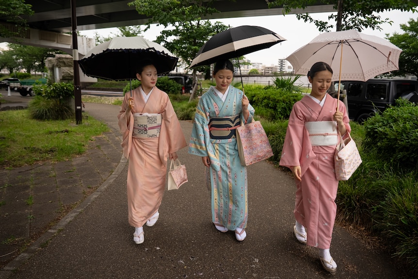 Three young woman walk holding umbrellas in kimonos.