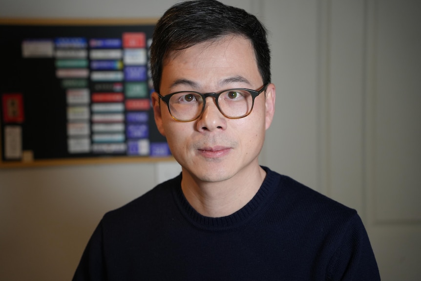 A dark-haired man in glasses in a room with an organisational board on the wall.