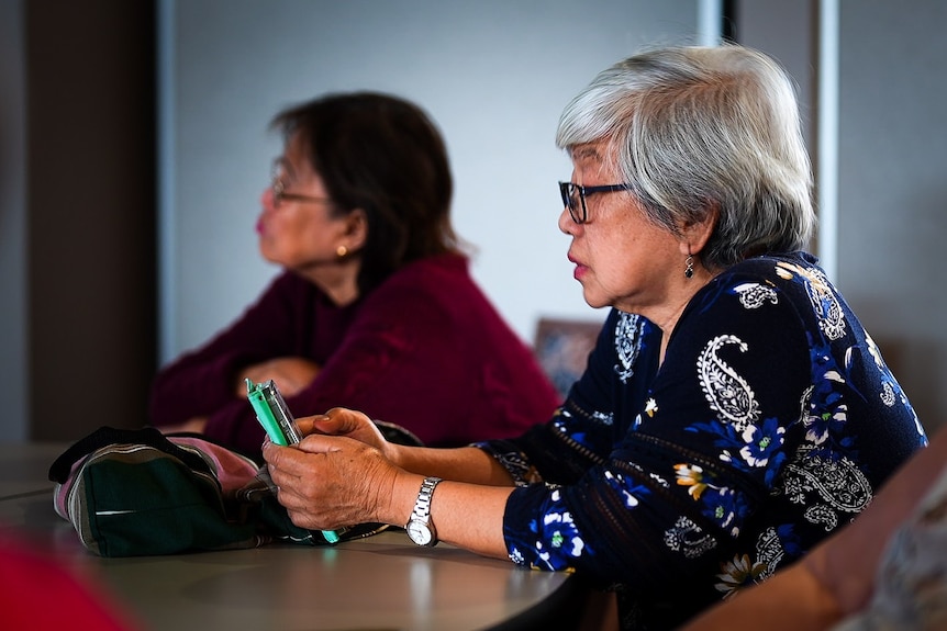 An older woman sitting at a table using a mobile phone. Another woman is in the background.