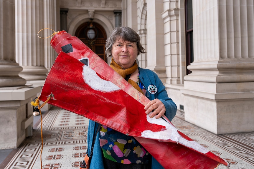 A grey haired woman in a blue coat holds a folded up red banner at Victorian Parliament.