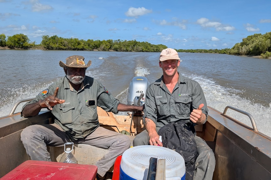 Two men siting on a mental speed boat smiling while thumbs upping the camera, both in green work shirts out at sea.