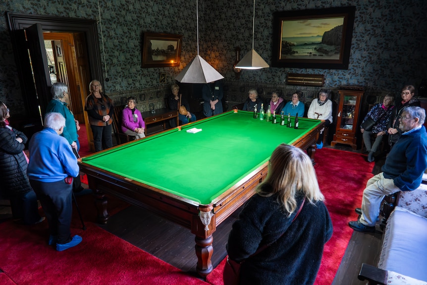A group gathers in large room around a full sized pool table inside a 19th century homestead listening to a man speak.