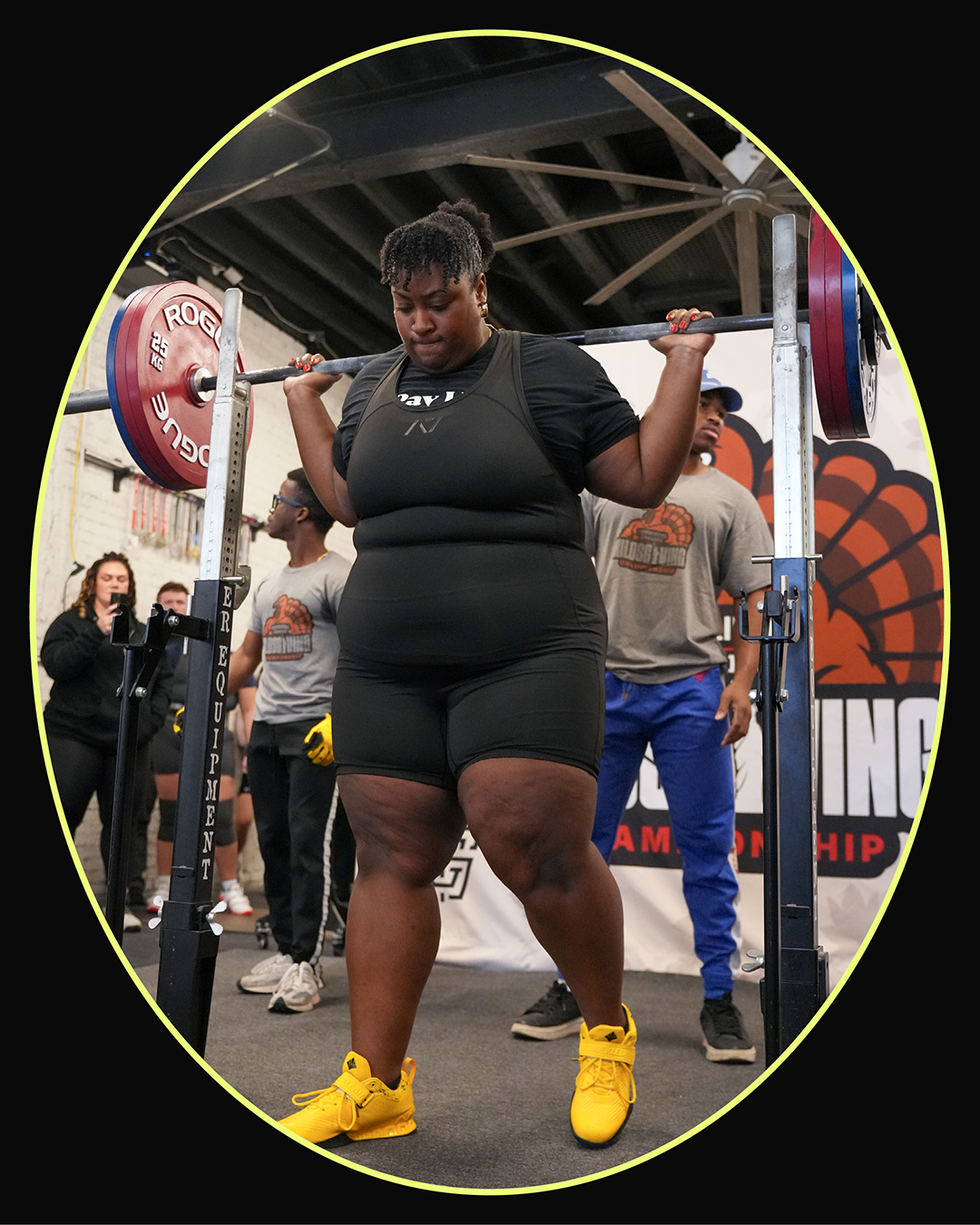 A weightlifter preparing to perform a squat lift at a powerlifting competition, with spectators in the background.