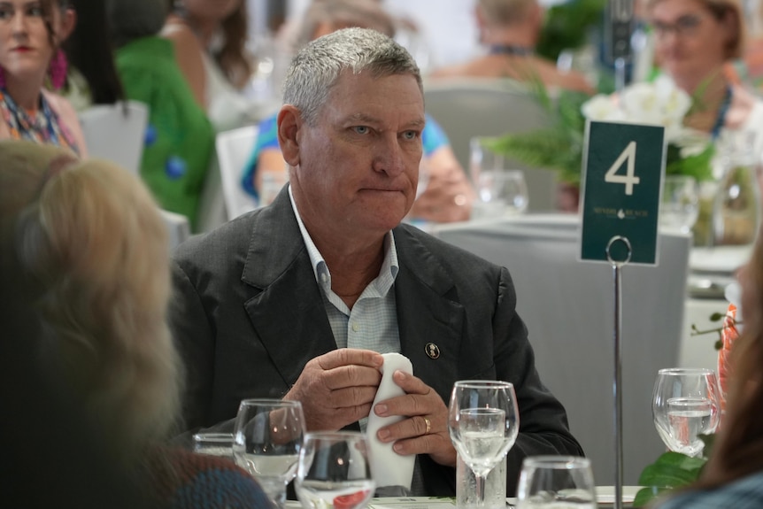 A man with short, grey hair sitting at a table for a luncheon event with a serious facial expression.