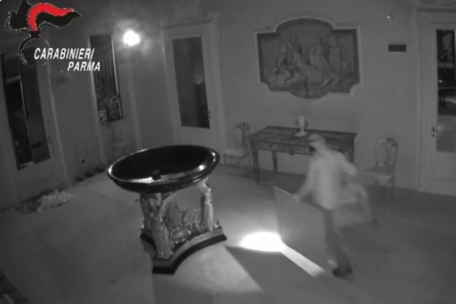 A masked figure in light clothing running through an open foyer area next to a large black basin while holding framed paintings.