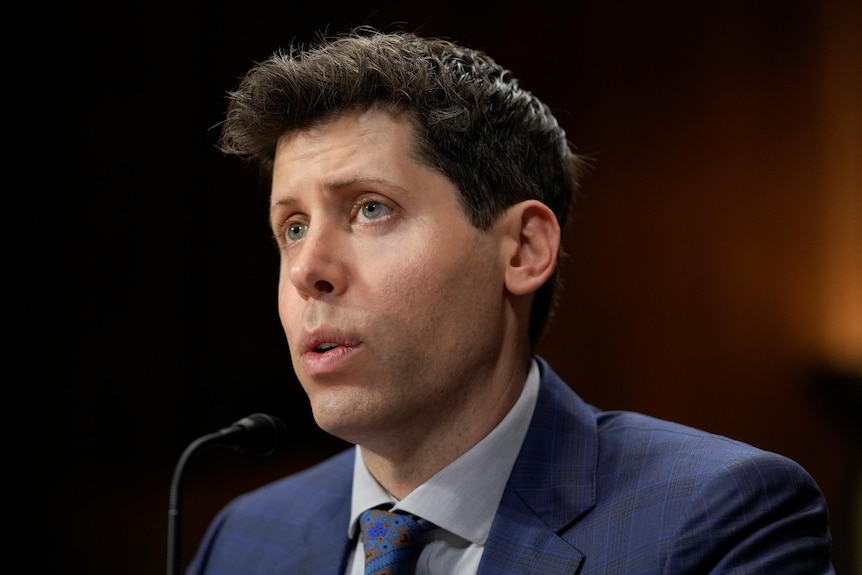 A white man with short hair speaks into a small desk microphone, while wearing a suit and tie