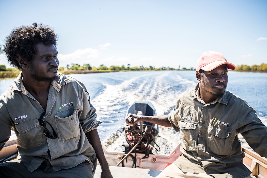 Two Aboriginal men on a speed boat travelling over ocean water, both looking right of frame.
