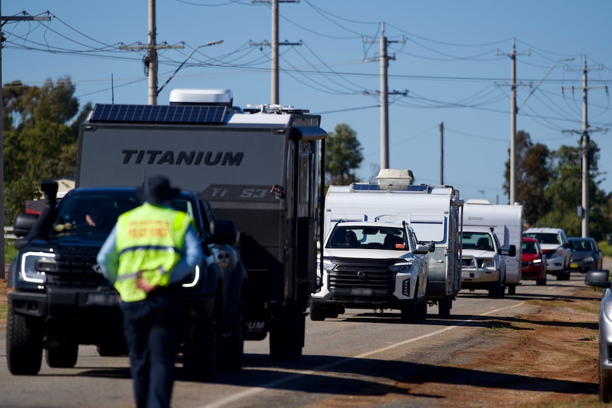 A man in a high vis vest walking towards a line of caravans on a road with power lines in the background.