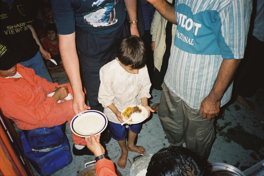 A young boy holds a plate of food, surrounded by adults.