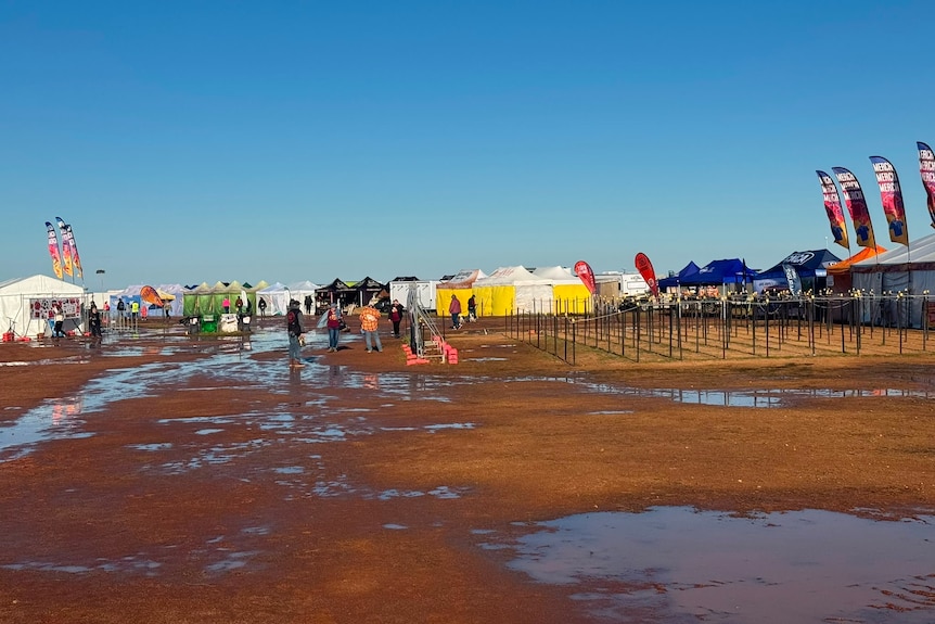 People walking around a claypan with water on it amongst a series of colourful tents and signs.