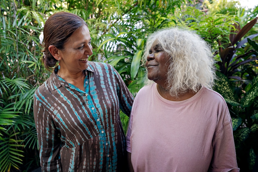 An Indian woman and an Indigenous woman smiling at one another, they are standing in front of a leafy garden.