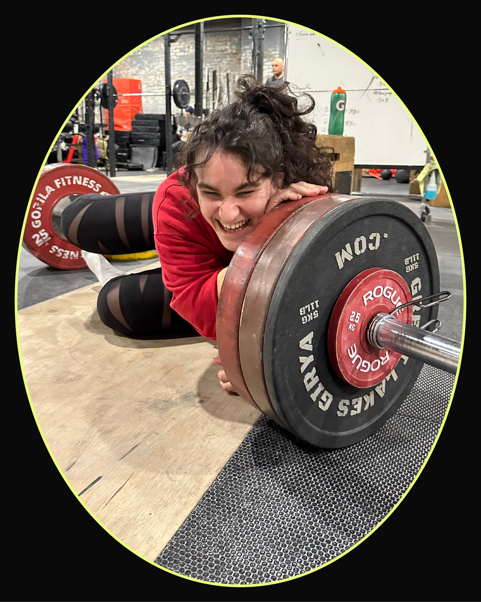A smiling woman enjoys lifting weights while resting her cheek on a barbell in a gym setting.