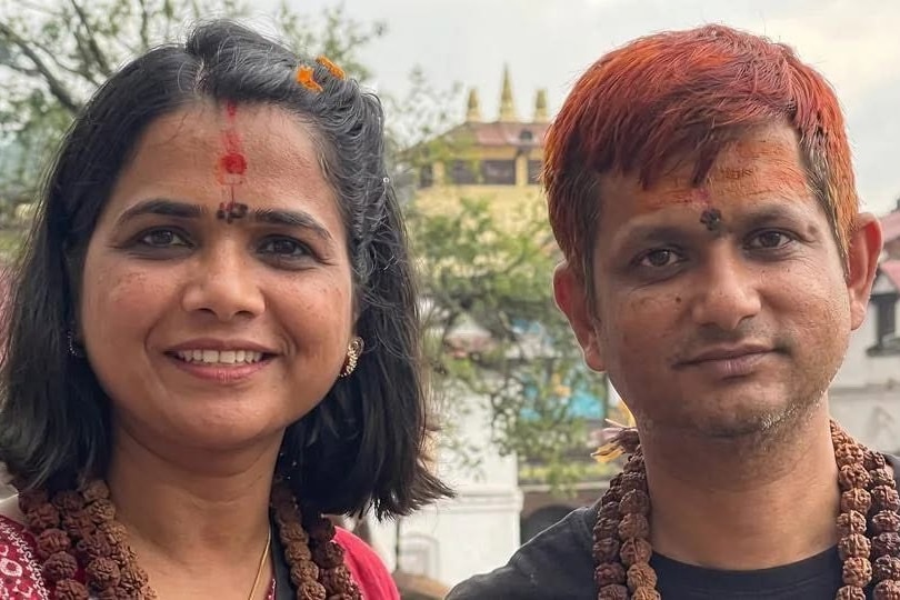 A dark-haired woman and a man with his hair dyed red wear beads as they stand in front of a temple.