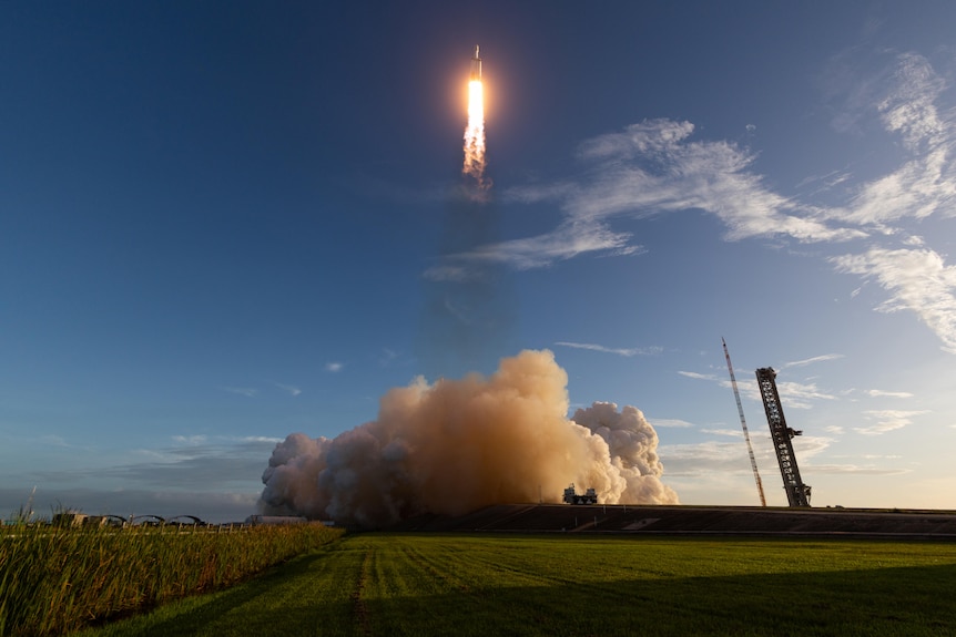 A bright rocket flying up from a cloud of launch dust in a sunny green field.