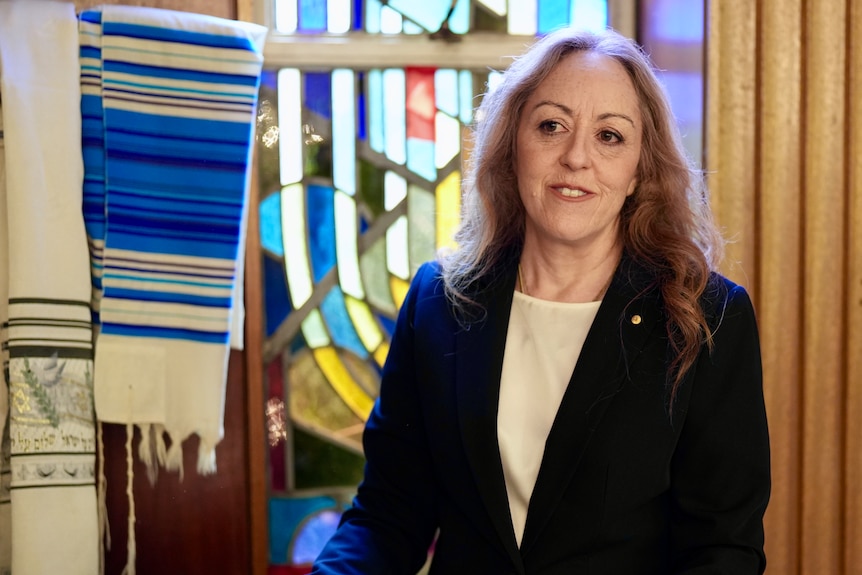 Rabbi Jacqueline Ninio standing inside a synagogue in a navy suit.