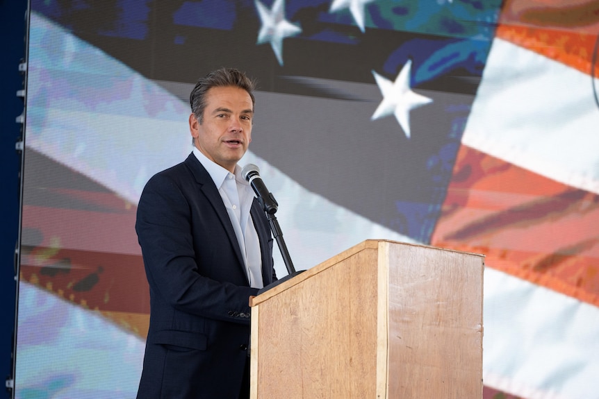 Lachlan Murdoch stands at a lectern to give a speech, the projection of a US flag behind on screen