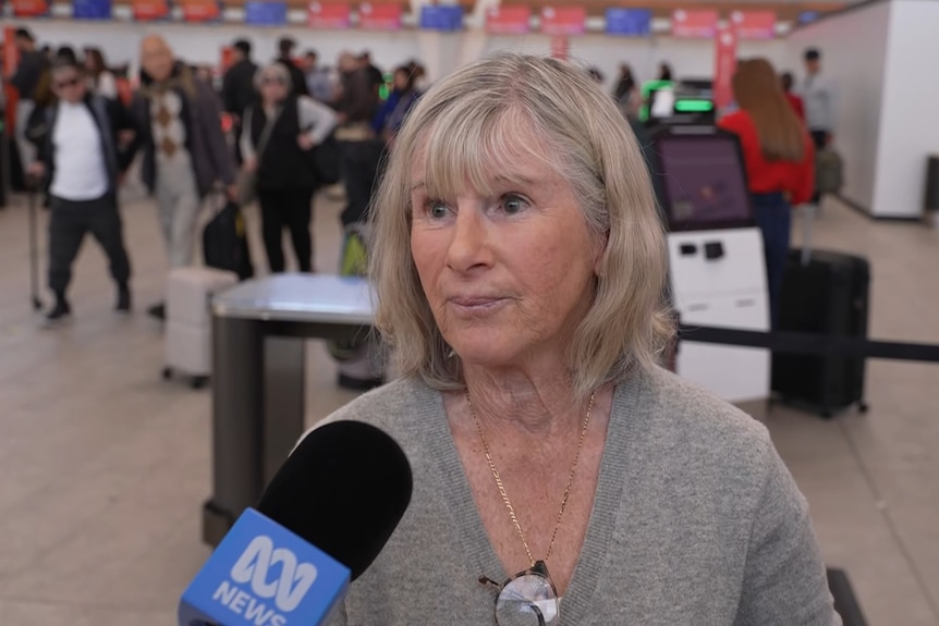 A woman with blonde/grey hair, in a grey vneck cardigan, speaking at the airport into an ABC microphone.
