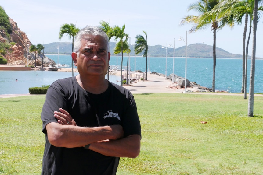 A man, looking stern, with his arms crossed in front of the beach.