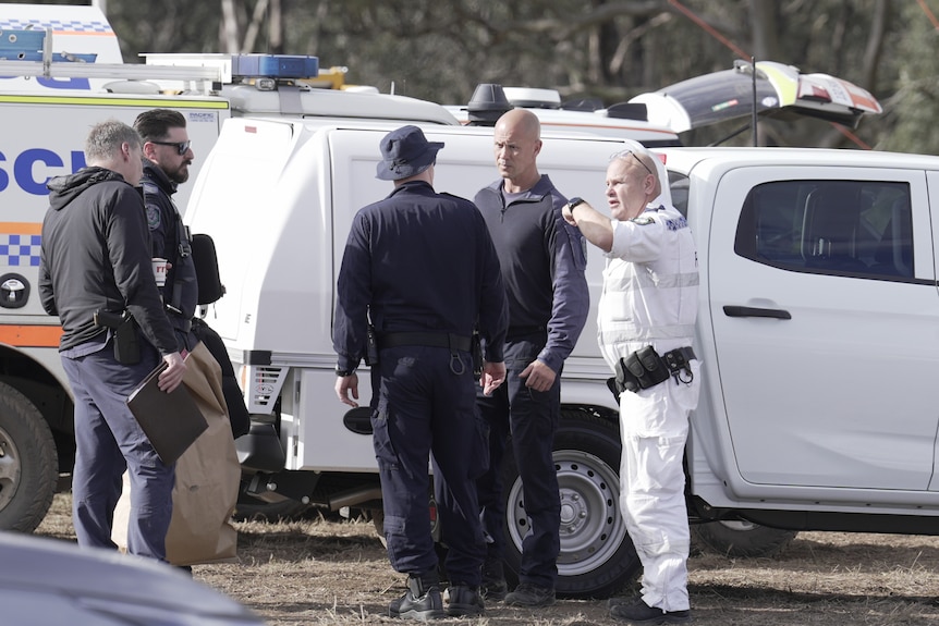 Bald man in white overalls gestures as he speaks with other police.