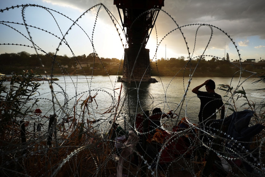 A silhouetted person stands behind loops of barbed wire on the banks of a river.