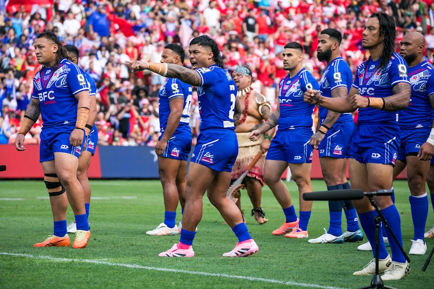 A group of Samoan men wearing blue rugby league uniforms stand on a field in a stadium challenging opposition