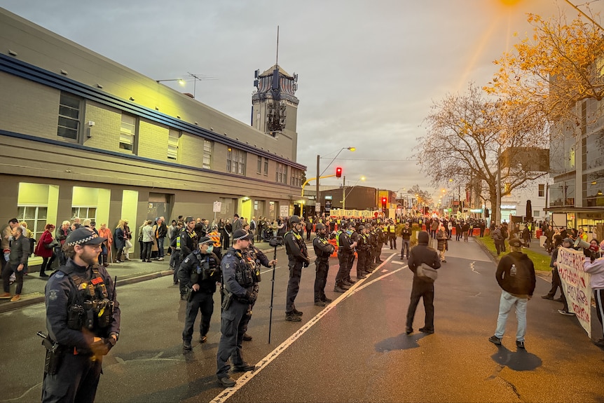 Police stand in a line in the middle of a road with a large group of people on the road in the distance.