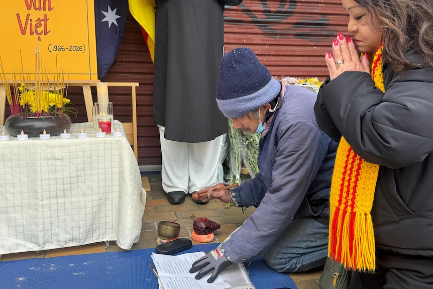A man in a blue jacket and beanie pends over a book on the ground while a woman in black kneels beside praying.