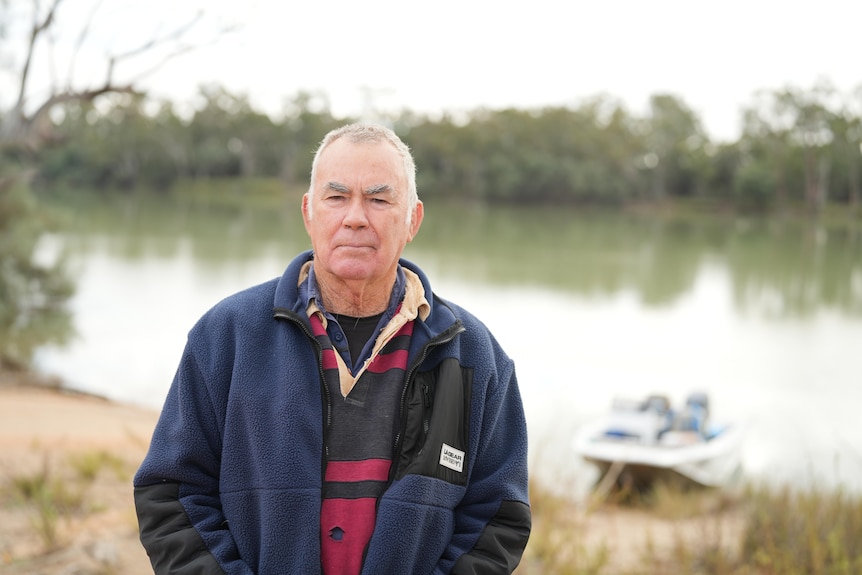  A fair-skinned older man in navy jacket and red and navy shirt in front of the River Murray. 