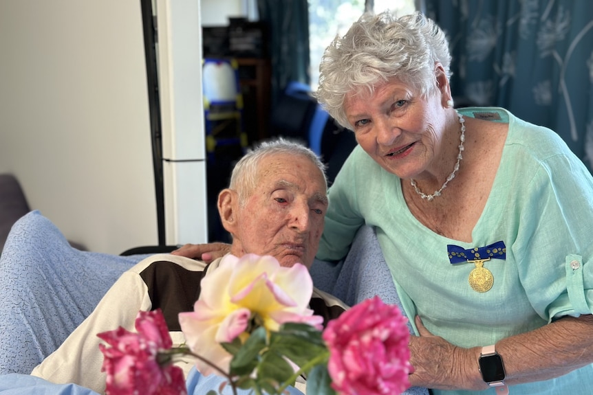 A woman wearing a medal stands closeby a frail man.
