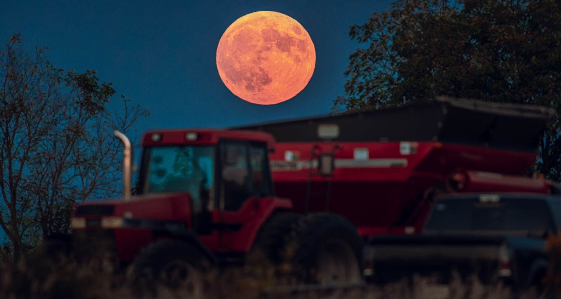 An orange full moon is photographed above a red tractor in a field at night.