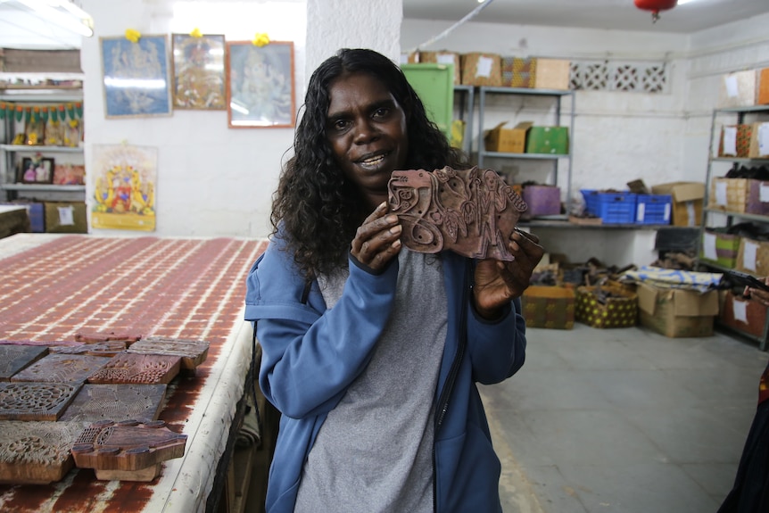 An Indigenous woman smiles are she holds up the wooden stamp her mother designed, she is standing inside a remote NT art studio.