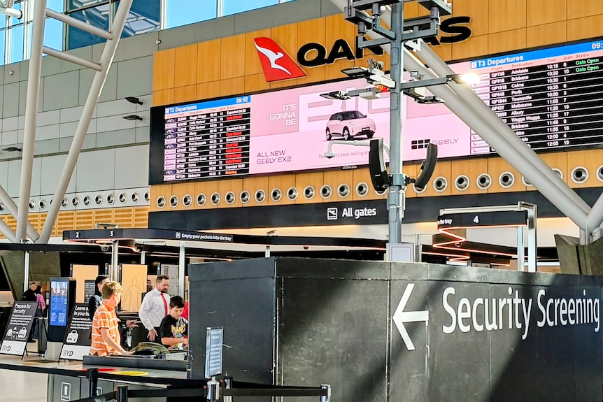 A flight arrival and depatures board in an airport, with a Qantas and Security Screening signs.