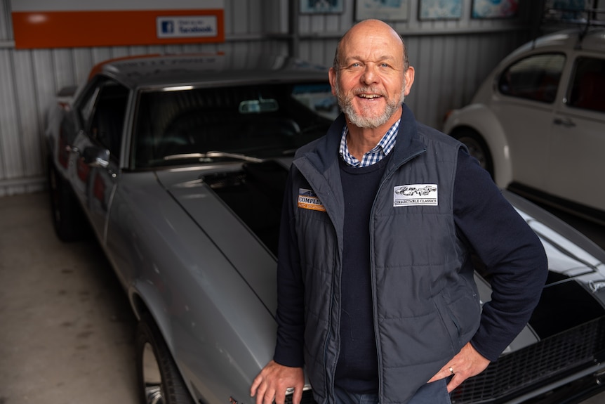 A man in navy jumper standing in front of a cool car, smiling at the camera.