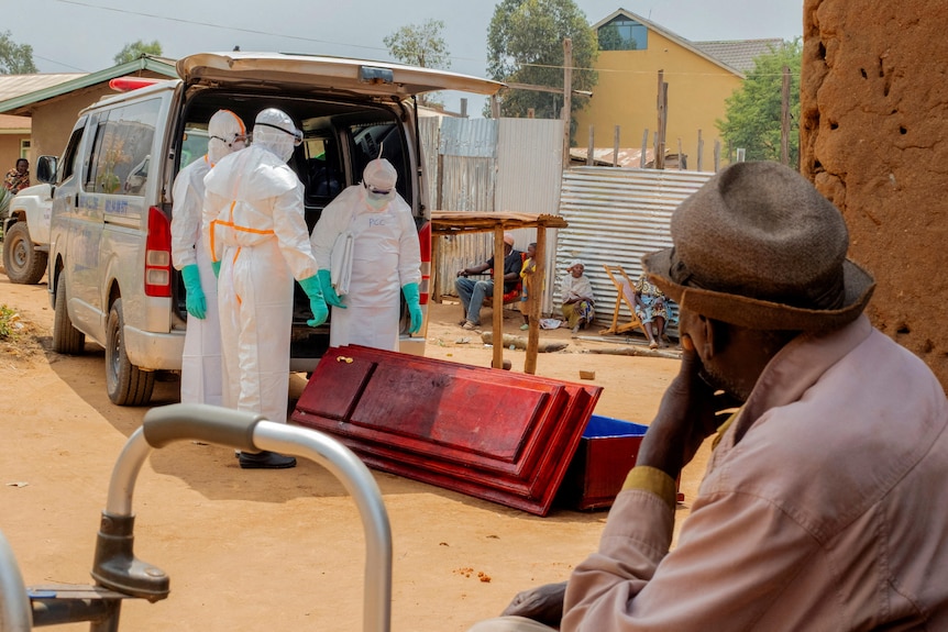A man wearing a hat sits by as three men in full body, white personal protective equipment stand around a coffin.